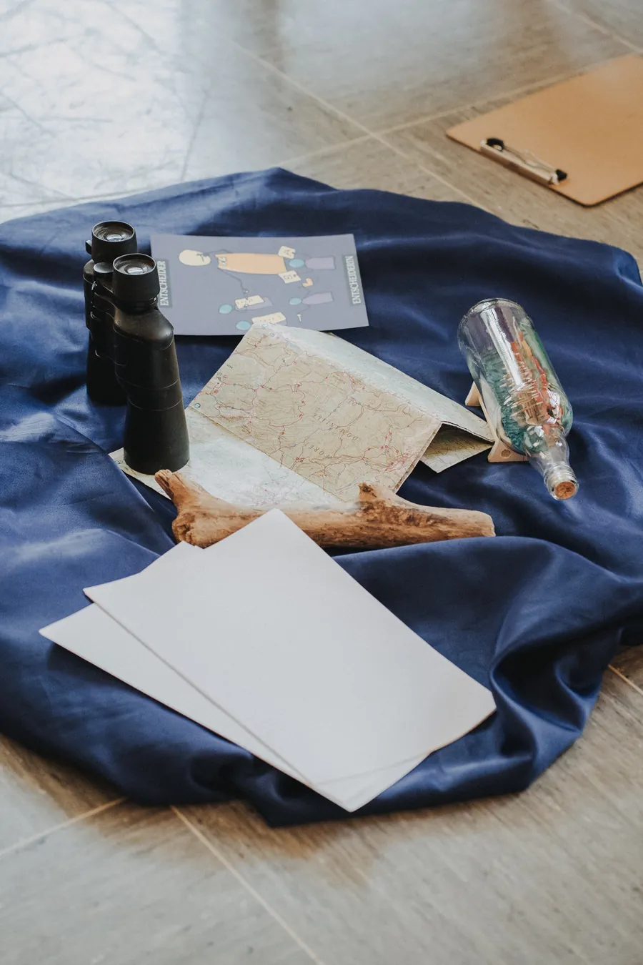 Fernglas, Landkarte, Flaschenpost und leere Papierbögen liegen auf einem blauen Tuch auf dem Boden