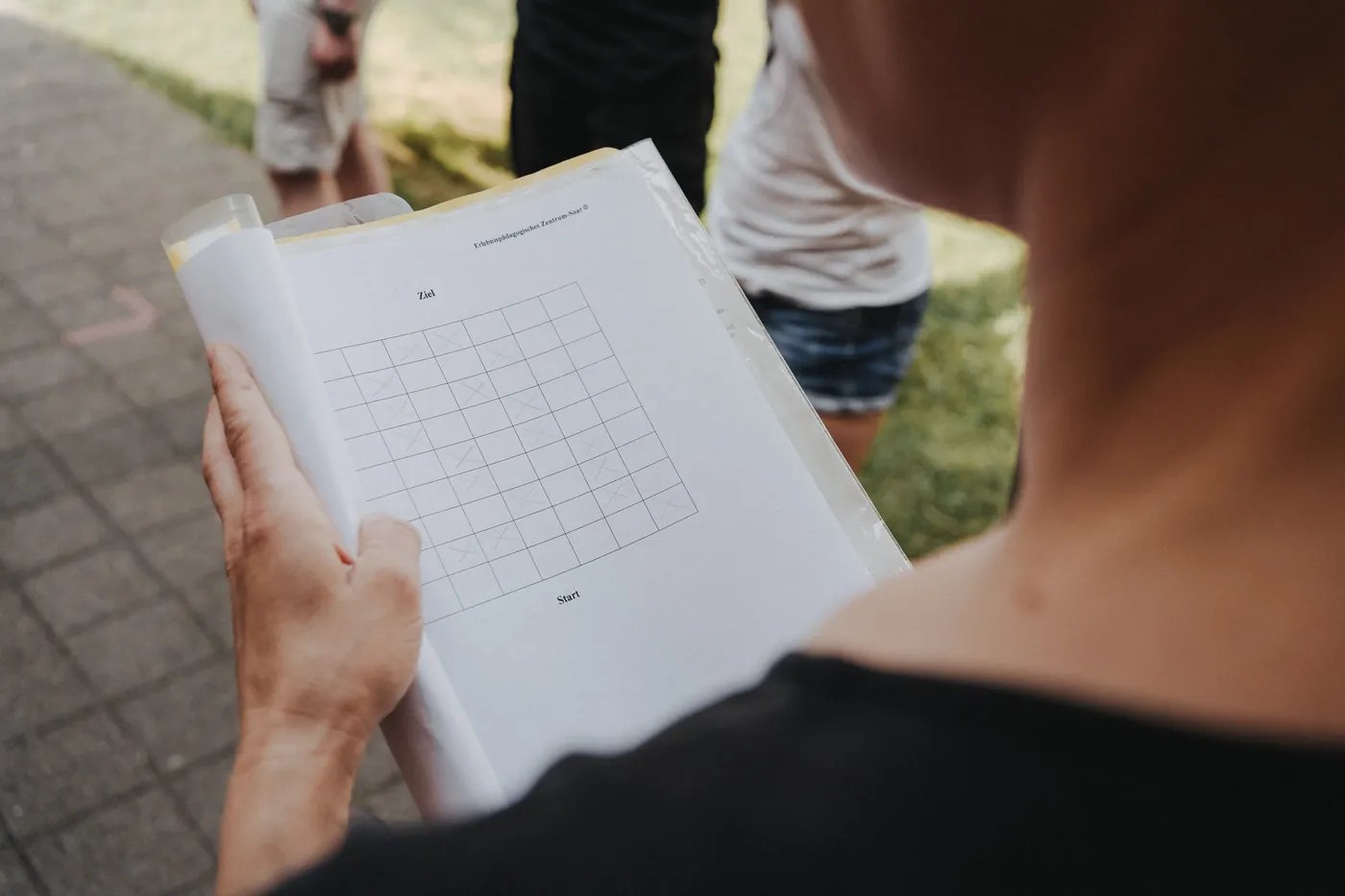 Eine Person hält eine Mappe mit einem Rasterplan, beschriftet mit Start und Ziel, im Hintergrund stehen Menschen im Freien